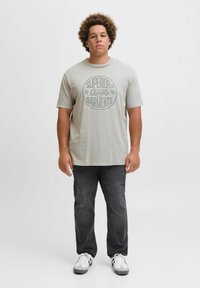 Hellgraues T-Shirt mit kurzen Ärmeln, das ein kreisförmiges Grafikdesign in dunklerem Grau aufweist. Getragen mit dunklen Jeans und weißen Sneakers mit schwarzen Akzenten.