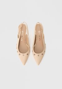 Zapatos planos slingback beige con punta puntiaguda y tachuelas decorativas metálicas a lo largo de los bordes, con la marca "GUESS" en la plantilla.