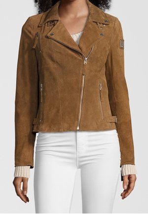 Veste de motard en daim marron avec fermeture éclair et détails de boutons portée sur un pull crème et un pantalon blanc, montrée sur une personne debout.