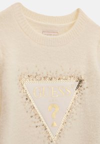 Pull couleur crème avec un logo triangulaire encadré de sequins dorés indiquant "GUESS USA" et un point d'interrogation doré à l'intérieur du triangle.