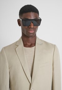 Blazer en lin beige avec un bouton unique, poche plaquée, associé à une chemise beige texturée et des lunettes de soleil noires surdimensionnées.