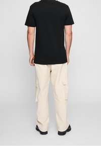 Černé bavlněné tričko s krátkým rukávem v kombinaci s béžovými cargo kalhotami, které mají boční kapsy. Kalhoty mají volný střih a jsou mírně pomačkané.