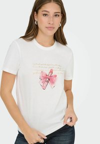 Jonge vrouw die een wit T-shirt draagt met een roze lippenstift en een strikillustratie en cursieve tekst, de zoom van het T-shirt vasthoudend, op een lichte achtergrond.