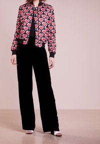 Woman wearing a black jacket with red pixelated hearts, black wide-leg pants, and white open-toe heeled sandals, standing on a beige floor.