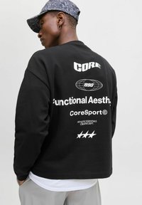 Hanorac negru cu text alb pe spate, având grafice și logo-uri. Împreună cu o șapcă întunecată cu model, plasată pe un fundal neutru.