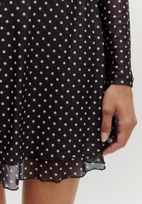Zwarte jurk met witte polka dots, lange doorschijnende mouwen en een gerimpelde zoom. Een hand is zichtbaar aan de rand van de jurk.