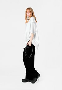 Camisa blanca de gran tamaño con un bolsillo, pantalones negros holgados con un detalle de cadena, y zapatos negros texturizados, todo sobre un fondo liso.