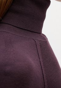 Gros plan d'une personne portant un pull à col roulé côtelé violet foncé avec des lignes de couture visibles sur l'épaule et le cou.