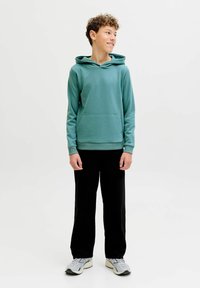 Dreng med krøllet hår iført en grønblå hættetrøje, sorte bukser og grå sneakers, der står og kigger til siden på en hvid baggrund.