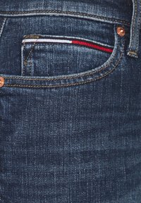 Vaqueros de mezclilla con un acabado azul oscuro, herrajes de cobre y un bolsillo frontal con un acento a rayas en rojo, blanco y azul.