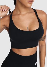 Žena v čiernej športovej podprsenke a legínach s vysokým pásom, drží jednu šnúrku podprsenky a ukazuje tetovania na hrudníku a ruke.