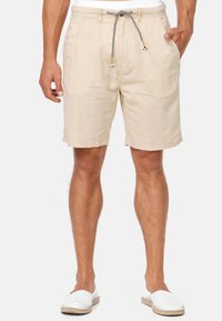 Man i beige linnebyxor med svart-vita dragsnören, vita slip-on-skor, och en hand i fickan mot vit bakgrund.