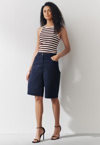 Débardeur rayé blanc et marron, associé à un short marine. Le short comprend une fermeture par bouton, des poches latérales et une coupe décontractée. Escarpins noirs.