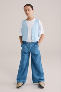 Helesinine varrukateta vest, millel viis nööpi, kantud valge T-särgi peale, koos laiade säärega teksapükste ja beežide kingadega.