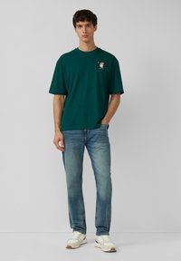 Young man standing against plain background, wearing dark green T-shirt with small Santa graphic and light blue jeans with white sneakers.