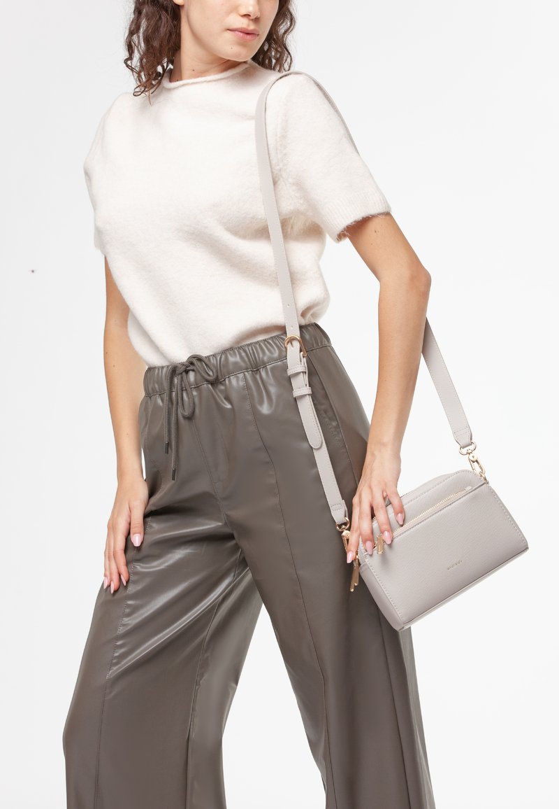 Maglione bianco morbido a maniche corte abbinato a pantaloni alti, grigio scuro e lucidi. Tiene una borsa a tracolla in pelle grigia chiara con dettagli dorati.