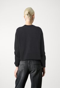 Svart kortermet hettegenser med lange ermer, laget av et mykt stoff, med rund hals og en avslappet passform. Brukt sammen med slitte grå jeans.