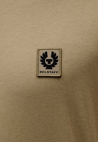 Χακί T-shirt από απαλό βαμβάκι, με τετράγωνο μαύρο έμβλημα με φτερά αετού και το κείμενο "BELSTAFF" στη  μέση του στήθους.