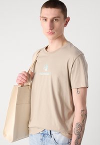 Šviesiai ruda medvilninė marškinėliai trumpomis rankovėmis su mėlynu "Calvin Klein Jeans" logotipu. Modelis laiko šviesiai rudą madingą rankinę. Reguliarus modelis.