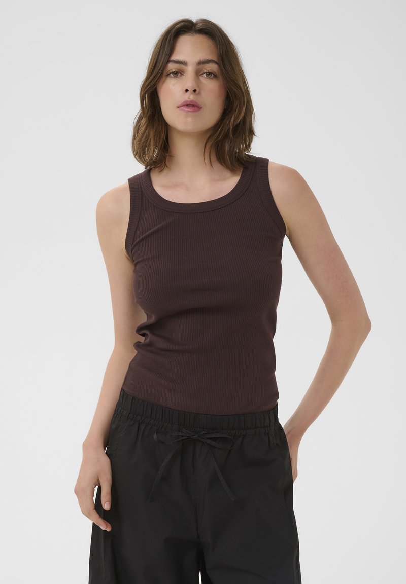 Donna con capelli castani fino alle spalle che indossa un top smanicato marrone scuro a coste e pantaloni neri con coulisse, in piedi davanti a uno sfondo semplice.