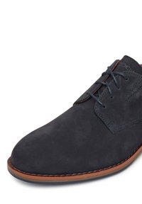 Granatowe zamszowe buty o zaokrąglonym czubku, z kontrastującymi brązowymi szwami i niebieskimi sznurowadłami. Zawierają teksturowaną cholewkę oraz płaską podeszwę.