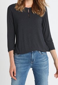 Svart blus med prickar och rund halsringning, nyckelhålsdetalj och trekvartsärmar, kombinerad med ljusblå jeans. Mjuk tygtextur.