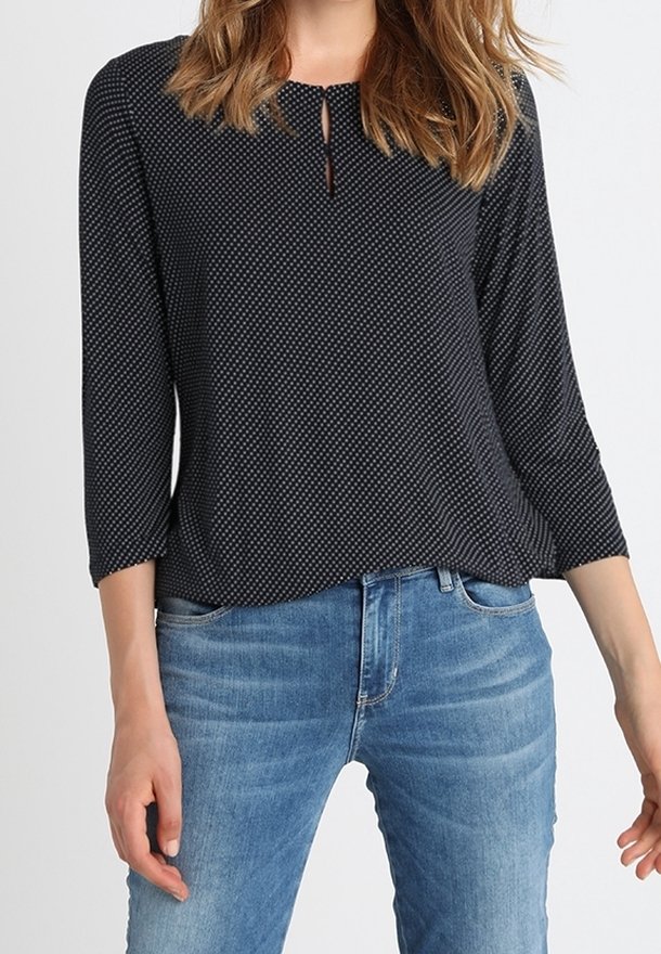 Svart blus med prickar och rund halsringning, nyckelhålsdetalj och trekvartsärmar, kombinerad med ljusblå jeans. Mjuk tygtextur.