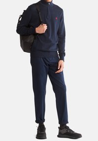 Suéter navy de medio cierre con puños acanalados, pantalones navy a juego y zapatos de senderismo negros, acompañado de una mochila negra.