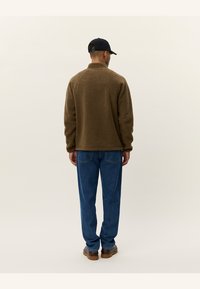 Brown fleece jacket with a high collar, paired with blue denim jeans. The outfit features a casual design and is worn with a black cap.