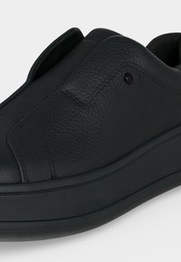 Μαύρο δερμάτινο παπούτσι χωρίς κορδόνια με υφή επάνω μέρος, στρογγυλεμένη μύτη και παχύ καουτσούκ πάτο, εμφανιζόμενο σε κοντινή λήψη.