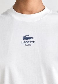 Balta medvilninė marškinė su apvaliu iškirpimu, su tamsiai mėlyna krokodilo logotipu ir užrašu "LACOSTE PARIS" spausdintu po logotipo.