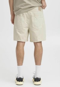 Beige Denim-Shorts mit geradem Schnitt, Fünf-Taschen-Design und einem Lederpatch am hinteren Bund. Getragen mit schwarzen Schuhen und weißen Socken.