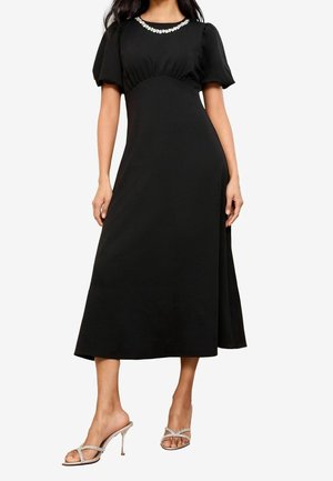 Robe midi noire avec manches bouffantes, corsage froncé et ornements décoratifs autour du décolleté. Portée avec des talons argentés à brides.