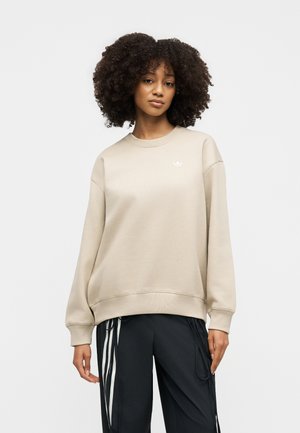 Žena s kovrčavom kosom koja nosi bež Adidas trenirku i crne hlače s bijelim prugama stoji na jednobojnoj svijetloj pozadini.