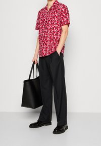 Czerwona koszula z krótkim rękawem z graficznym wzorem, w połączeniu z czarnymi szerokimi spodniami i błyszczącymi czarnymi butami, trzymająca czarną torbę na ramię.