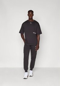 T-shirt preta oversized de manga curta com logótipo branco e calças de fato de treino pretas afuniladas, complementadas por ténis desportivos brancos.