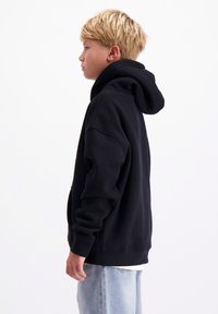 Zwart hoodie gemaakt van zacht materiaal, met een voorkant zak, ribgebreide boorden en zoom. Getoond op een model in profiel tegen een effen achtergrond.
