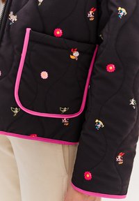 Čierna prešívaná bunda s ružovým lemom a veľkou kapsou, zdobená farebnými vzormi postáv a kvetov. Mäkká textúra, ležérny dizajn.