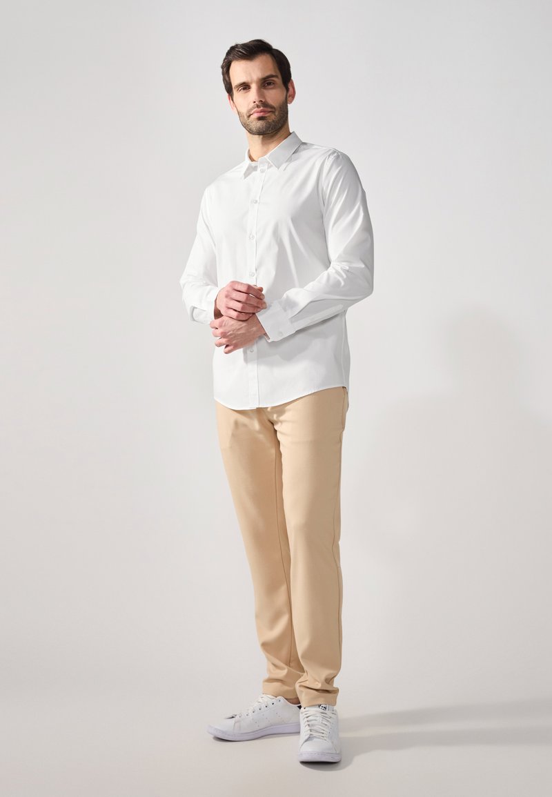 Weißes Langarmhemd mit einem Button-Down-Kragen, kombiniert mit beigen Hosen und weißen Sneakers, dargestellt an einem Mann, der mit verschränkten Armen dasteht.
