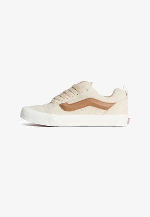 Beżowy zamszowy sneaker z brązowymi akcentami, białą płaską podeszwą, okrągłym noskiem i sześcioma dziurkami na sznurowadła. Posiada małe wycięcia po bokach.