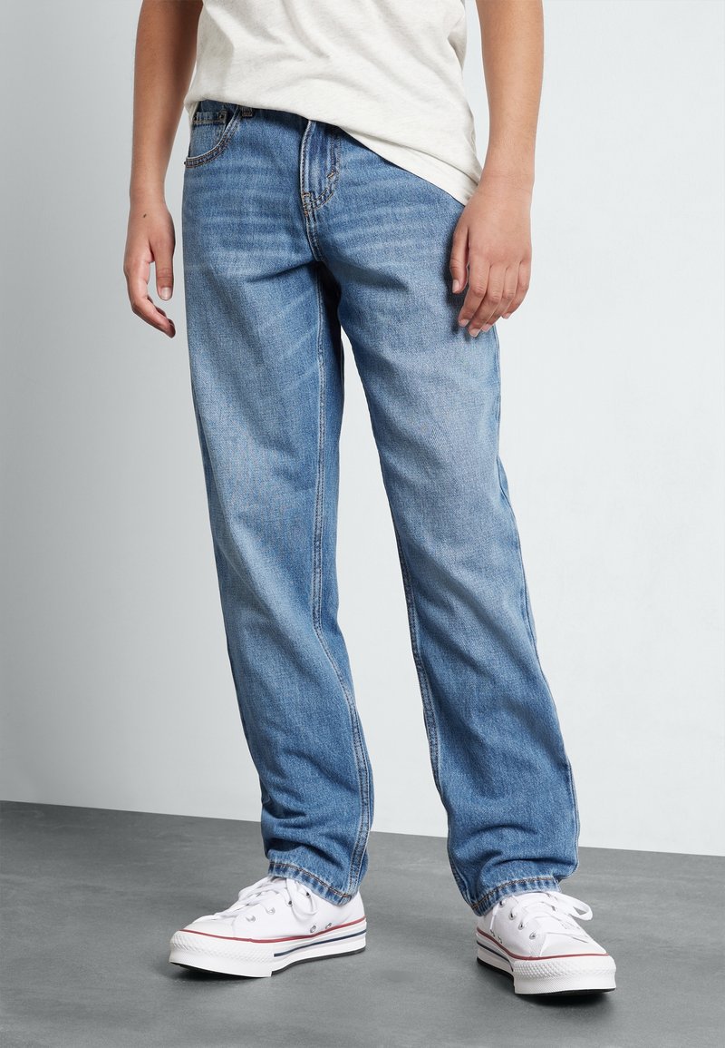 Personne portant un jean droit bleu clair et des baskets blanches, debout sur un sol gris devant un mur blanc.