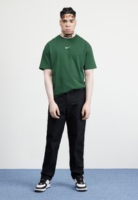 Grön t-shirt med kort ärm och Nike-logotyp, kombinerad med svarta byxor och svartvita sneakers, mot en ljus bakgrund.