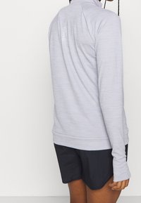 Top a maniche lunghe grigio chiaro con una finitura testurizzata, colletto alto e aperture per il pollice; abbinato a pantaloni corti neri. Design semplice e funzionale.