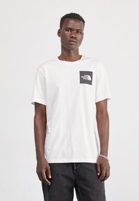 Bijela pamučna majica s kratkim rukavima, s crnim kvadratnim džepom na prsima s logom "The North Face". Model nosi tamne hlače.