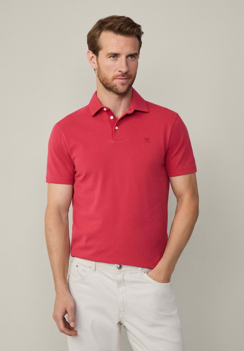 Červená polokošeľa vyrobená z bavlny, s klasickým golierom, trojbuttonovým zapínaním a vyšitou logom na hrudi. Nosená s bielymi nohavicami.