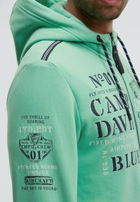 Sudadera verde menta con cremallera, con gráficos impresos en blanco y negro. Tiene una tela texturizada y acentos de rayas en contraste.