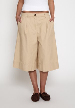 Frau trägt beige knielange weite Shorts, weißes kurzes Top und braune Loafers, steht mit Händen in den Taschen.