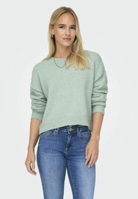Pull en maille texturée vert clair avec un col rond et des épaules tombantes, associé à un jean en denim bleu. Design simple et court.