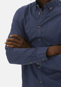 Camisa abotonada azul marino con cuello puntiagudo, mangas largas, bolsillo en el pecho y botones en tono dorado. Tejido suave y corte clásico.
