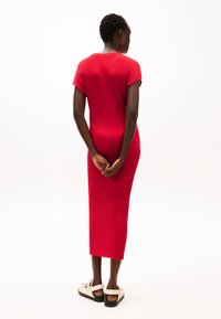 Abito a maglia rosso a coste con maniche corte, design aderente e lunghezza media. Il modello sta in piedi con le mani dietro la schiena, indossando sandali di colore chiaro.